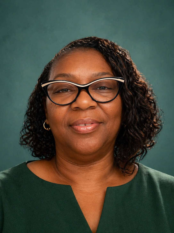 A professional portrait of a woman with curly dark hair wearing cat-eye glasses with a white and black frame, a dark green top, and small gold hoop earrings, set against a solid dark green studio background.